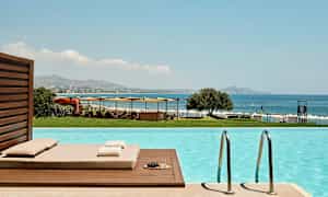 Udsigt fra en pool mod havet, liggestole og parasoller. (AI-genereret tekst)