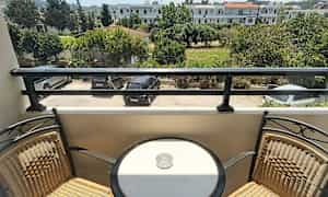 En altan med to stole og et rundt bord med et askebæger, med udsigt over en parkeringsplads og grønt område. (AI-genereret tekst)