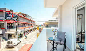 En altan med bord, stol og blomster i forgrunden samt udsigt over en gade med butikker og bygninger. (AI-genereret tekst)