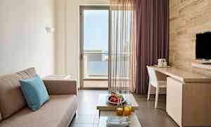 Et værelse med en beige sofa, et bord med frugt og juice samt en altandør med udsigt. (AI-genereret tekst)