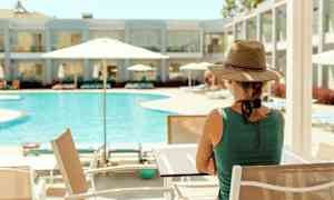 En person sidder ved et bord nær en pool med parasoller. I baggrunden ses en hotelbygning. (AI-genereret tekst)