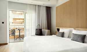 Billede af et hotelværelse med stor seng og siddeområde ved vinduet, der fører ud til altanen. (AI-genereret tekst)