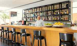 En moderne bar med høje stole ved en bardisk i træ, hylder fyldt med flasker og frugtkurve med citrusfrugter. (AI-genereret tekst)