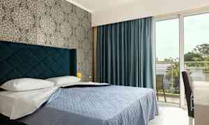 Et hotelværelse med en seng, blå sengetæppe og blå gardiner, samt et vindue, der fører til en altan. (AI-genereret tekst)