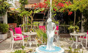 En fontæne med en hvid statue af en kvinde i en frodig have med borde og stole. (AI-genereret tekst)