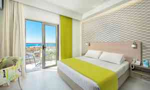 Et hotelværelse med en seng, grønt sengetæppe, lyse møbler og en altan med havudsigt. (AI-genereret tekst)