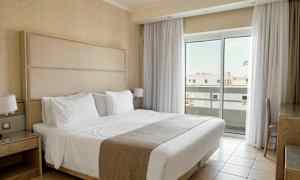 Et hotelværelse med en dobbeltseng beklædt med hvide sengelinned, natborde og store vinduer med gardiner, der lukker lys ind. (AI-genereret tekst)