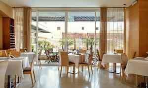 Et moderne rum med opdækkede borde og stole samt store vinduer mod en gårdhave med træer og møbler. (AI-genereret tekst)