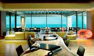 Et moderne rum med store glasvinduer, der viser udsigt over havet. Der er et langt træbord i forgrunden og en siddegruppe med moderne design i baggrunden. (AI-genereret tekst)