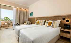 Et hotelværelse med to enkeltsenge, en siddegruppe og en altan med udsigt til grønt og himmel. (AI-genereret tekst)