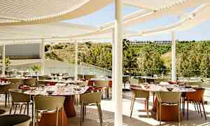 En udendørs restaurant med spiseborde og stole under en moderne hvid solafskærmning. Baggrunden viser grønne områder og bakker. (AI-genereret tekst)