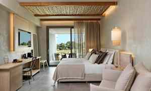 Et moderne hotelværelse med en dobbeltseng, en siddegruppe og et skrivebord. Gardiner lader dagslys komme ind fra altandøren. (AI-genereret tekst)