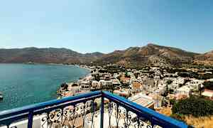 Udsigt fra en altan med blå gelænder over en kystby med hvide bygninger og omkringliggende bjerge. (AI-genereret tekst)