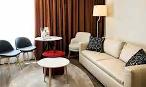 En stue med en lys sofa, et rundt bord, to stole og brune gardiner. (AI-genereret tekst)