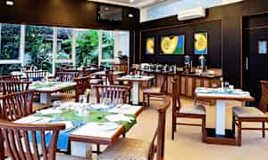 En restaurant med træborde og stole, farverige bordduge og dekorationer. Baggrunden viser en buffet med serveringscontainere. Vinduer giver udsigt til grønne planter. (AI-genereret tekst)