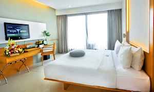 Et moderne hotelværelse med en stor komfortabel seng, skrivebord med blomsterarrangement og et TV på væggen. Vinduer med lyse gardiner lukker dagslys ind. (AI-genereret tekst)