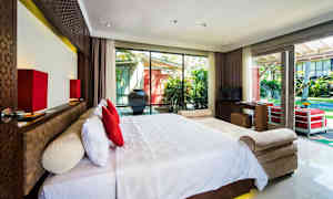 Et hotelværelse med en stor seng med hvidt sengetøj og røde puder foran et stort vindue, der vender ud mod en have. (AI-genereret tekst)