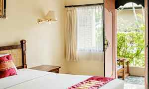 Et lyst hotelværelse med en seng dækket af hvidt sengetøj og en dekorativ rød løber. Et vindue med gardiner og en åben dør fører til en terrasse med grønne områder. (AI-genereret tekst)