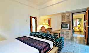 Et hotelværelse med en stor seng, en lille siddegruppe, et skrivebord med spejl og et gammelt fjernsyn. Badeværelset er til venstre, og en forbindelse til et andet rum ses i baggrunden. (AI-genereret tekst)