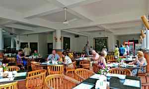 Et restaurantområde med borde og stole, hvor folk sidder og spiser. Indretningen er lys med hvide vægge og loft. (AI-genereret tekst)