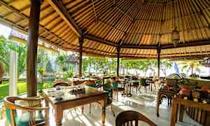 En udendørs restaurant med træborde og stole under et stråtag, omgivet af grønne planter og placeret tæt på stranden. (AI-genereret tekst)