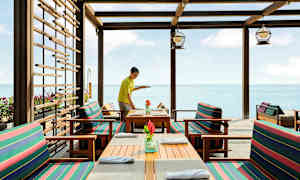 Restaurant ved havet med træborde og stole med stribede puder. (AI-genereret tekst)