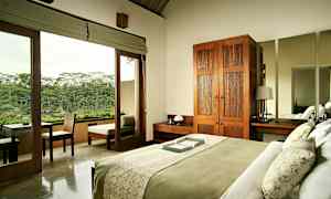 Et soveværelse med en seng opredt med grønt og hvidt sengetøj. Rummet har et træskab, natbord og et stort spejl. Glasdøre fører ud til en balkon med udsigt over frodig natur. (AI-genereret tekst)