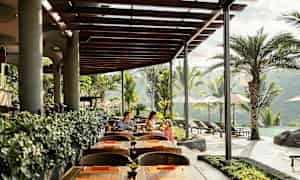 En udendørs restaurant med træborde og stole, træplanketag og grønne planter. En pool og palmer ses i baggrunden. (AI-genereret tekst)