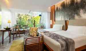 Et hotelværelse med to senge, et siddeområde og store vinduer, der lukker lys ind og giver udsigt til grønne træer. (AI-genereret tekst)