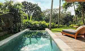 Billede af en pool med klart vand omgivet af frodig vegetation og en liggestol på en træterrasse. (AI-genereret tekst)