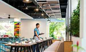 En restaurantlokale med borde og stole, hvor en person bærer en bakke med kopper. (AI-genereret tekst)