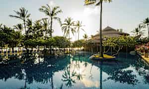 En pool omgivet af palmer og vegetation med en hytte med stråtag i baggrunden. (AI-genereret tekst)