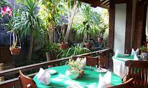 Et udendørsområde med borde og stole på en terrasse, dekoreret med grønne duge, tallerkener og servietter. Baggrunden domineres af grøn vegetation og hængende planter. (AI-genereret tekst)