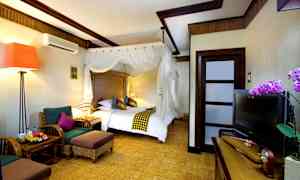 Et hotelværelse med en stor seng med en hvid baldakin, siddeområde med to lænestole og et bord, et TV på en skænk og en orkidédekoration ved siden af. (AI-genereret tekst)