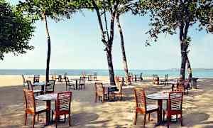 Udendørs borde og træstole placeret på en sandstrand med havet i baggrunden under træer. (AI-genereret tekst)