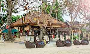 En strandrestaurant med stråtag, omgivet af træer og siddepladser i flettet rattanmøbler. (AI-genereret tekst)