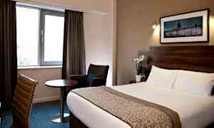 Hotelværelse med seng, stol, bord og vindue med gardiner. (AI-genereret tekst)