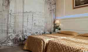 Et hotelværelse med to senge dækket med guldfarvede sengetæpper. Baggrundsvæggen har et kunstværk med arkitektoniske søjler og bygninger. En natlampe og en plante står på natbordet. (AI-genereret tekst)