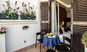 En balkon med et lille bord dækket til morgenmad og to stole. Bordet er dækket med en blå dug og fører ind til et soveværelse. (AI-genereret tekst)