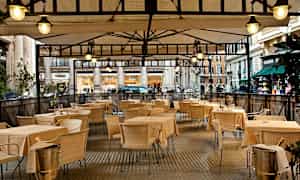 Billede af en udendørs serveringsområde med borde og stole under et overdækket tag. (AI-genereret tekst)