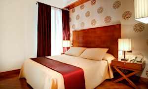 Soveværelse med en stor seng dækket med beige og vinrødt sengetøj, to sideborde med lamper og dekoreret tapet på væggen. (AI-genereret tekst)