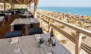 En restaurant med dækkede borde tæt på en strand med parasoller og liggestole. (AI-genereret tekst)