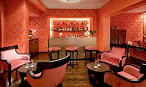 Billede der viser et elegant område indrettet som en cocktailbar med mørke møbler, lyserøde puder og flasker på en hylde. (AI-genereret tekst)