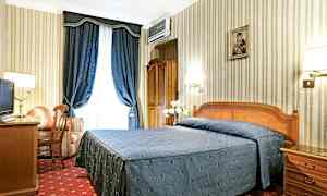 Et hotelværelse med en dobbeltseng dækket med et blåt sengetæppe, et skrivebord, en lænestol og blå gardiner. (AI-genereret tekst)