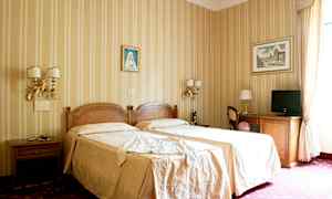 Et hotelværelse med to enkeltsenge dækket med beige sengetøj, dekorerede vægge med malerier og møbler inklusive borde, stole og et fjernsyn. (AI-genereret tekst)