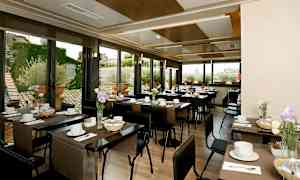 En lys restaurant med dækkede borde, dekoreret med blomster og vinduer der viser en udendørsmiljø. (AI-genereret tekst)