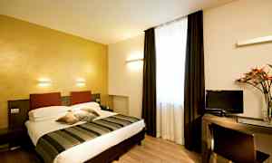 Et hotelværelse med en dobbeltseng, et skrivebord og et fjernsyn. Værelset har lyse vægge og et stort vindue med mørke gardiner. (AI-genereret tekst)