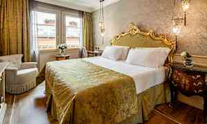 Et elegant soveværelse med en udsmykket seng med hovedgærde, dækket med et gyldent sengetæppe. Rummet har to vinduer og et bord med en vase med blomster. (AI-genereret tekst)