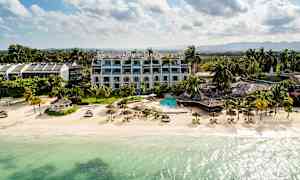 Strand med klart vand, parasoller og liggestole, omgivet af hotel og grønne områder. (AI-genereret tekst)