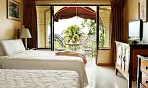 Et hotelværelse med to enkeltsenge, et fjernsyn på en trækommode og en åbning til en altan med udsigt til vegetation og palmetræer. (AI-genereret tekst)